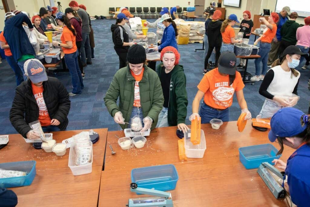 An overview of the room, student volunteers weigh each bagged meal on a scale to insure it's within an acceptable range.