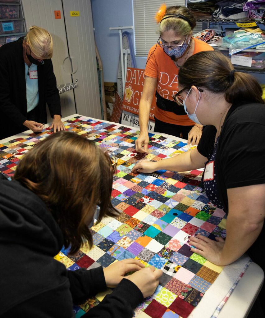 Event organizer Radha Selvester, in orange shirt, leads volunteers as they pin the top and bottom of a quilt to batting, which will fit between them and provide insulation.