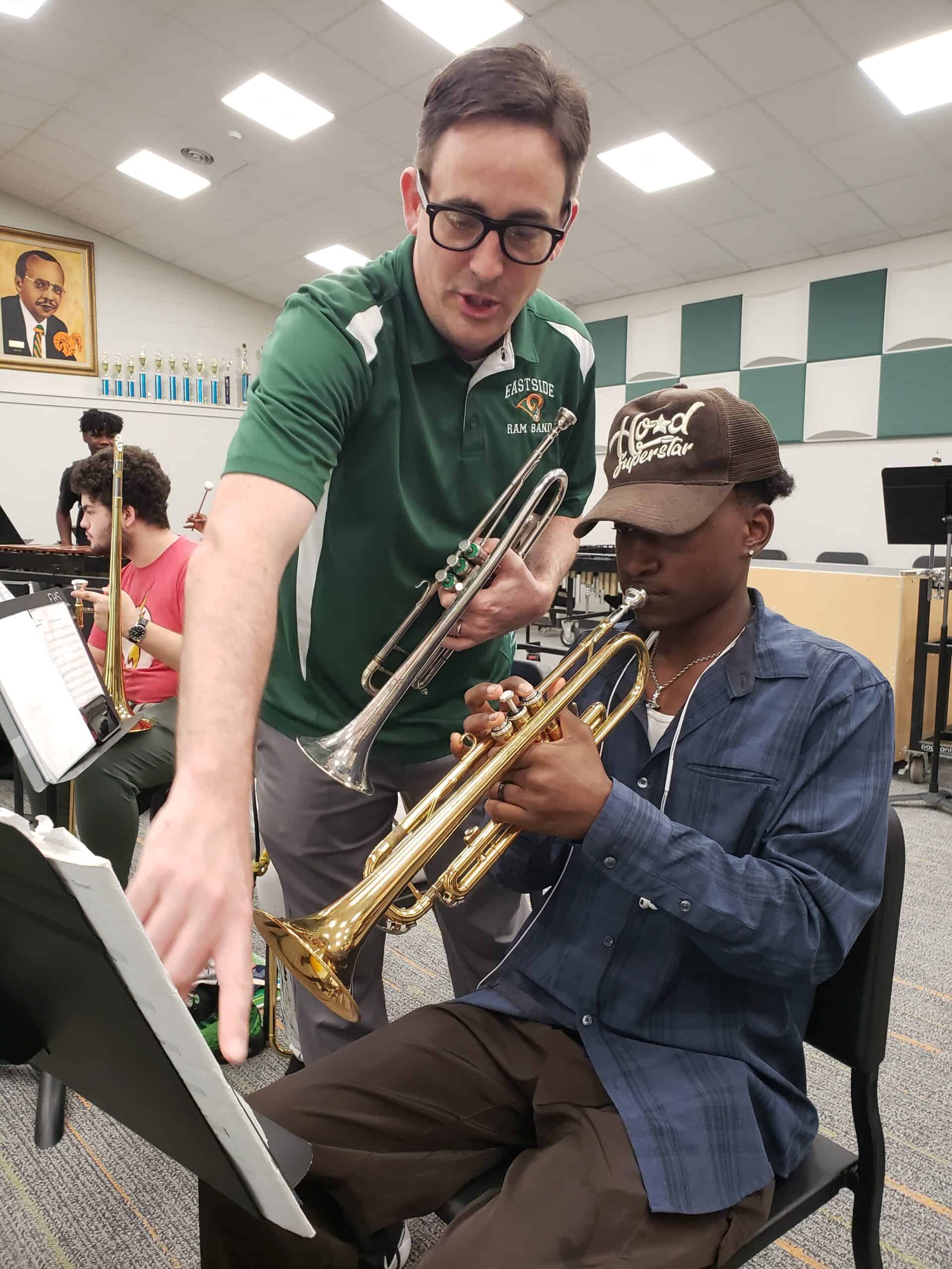 A man holding a trumpet stands and points to sheet music on a stand while a teen holding a trumpet to his mouth fingers the notes on the trumpet.