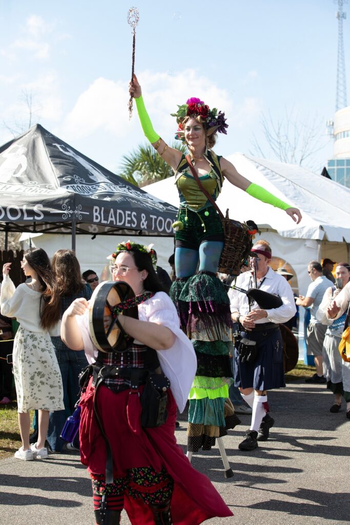 A woman balancing on stilts waves a wand to make bubbles.