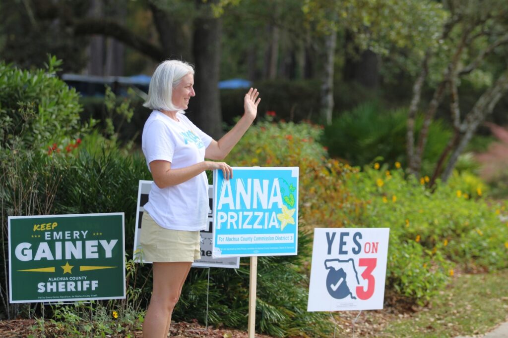 Alachua County Commissioner Anna Prizzia waves at vehicles along NW 34th Street at Westside Park, the location of precincts 26 and 32. 