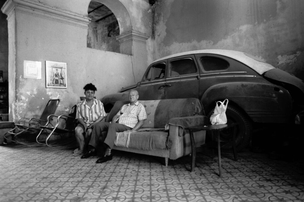 Randy Batista’s “El Tesoro de la Sala (The Living Room Treasure).” Havana, 1996, depicts a Cuban family keeping their car indoors to prevent thieves from stealing parts.