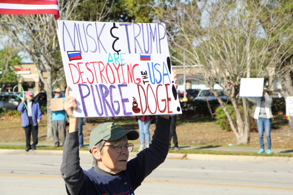 Protester Chris stands in front of Gainesville's Tesla dealership displaying a sign for drivers on North Main Street.