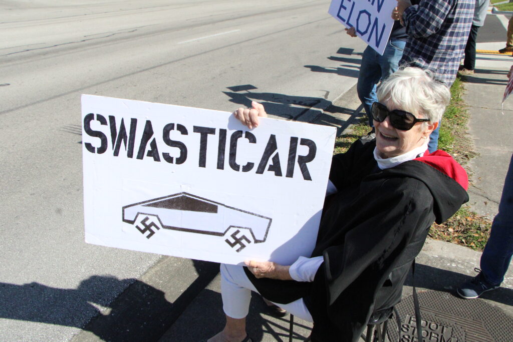 Protester Maggie protests Elon Musk and Tesla in front of Tesla dealership.