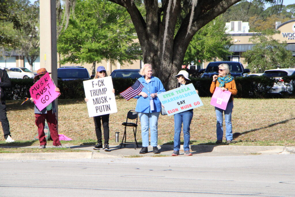 Tesla Takedown protesters hold signs across from the Tesla dealership in Gainesville.